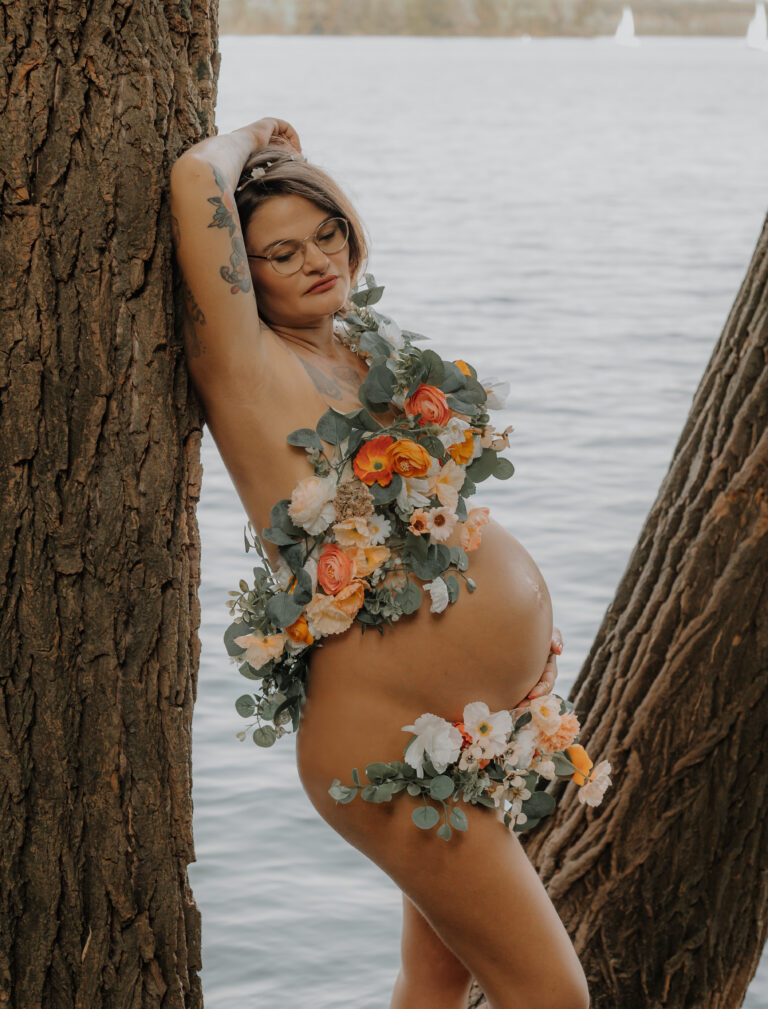 Schwangere mit Blumenschmuck lehnt an einem Baum am See